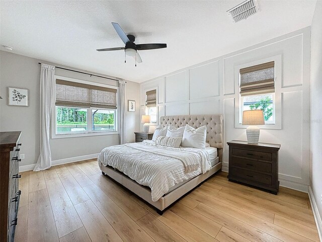 bedroom featuring a ceiling fan, visible vents, baseboards, light wood-style floors, and a decorative wall