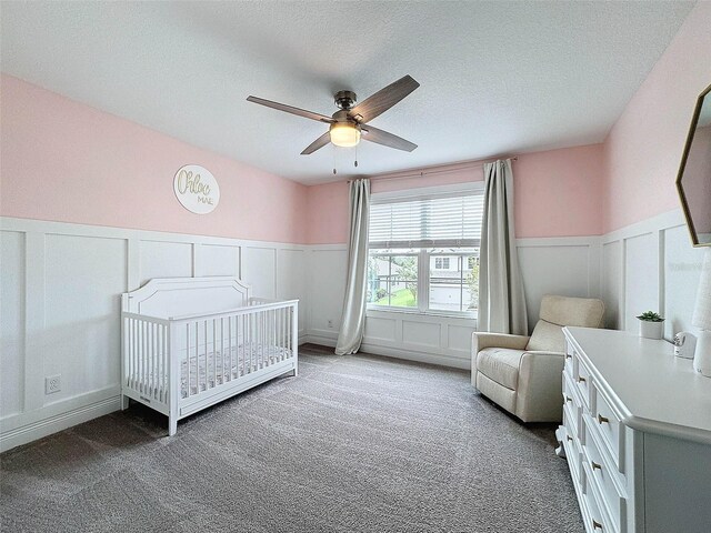 carpeted bedroom featuring a wainscoted wall, a nursery area, a ceiling fan, and a textured ceiling