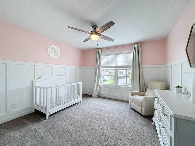 carpeted bedroom featuring a ceiling fan, a wainscoted wall, a nursery area, and a textured ceiling