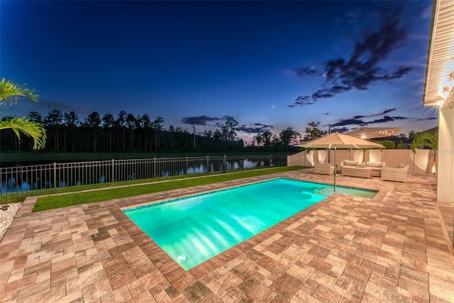 pool at dusk with an outdoor living space, a water view, a fenced backyard, and a patio