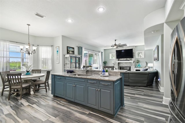 kitchen featuring stainless steel fridge, sink, a stone fireplace, a kitchen island with sink, and ceiling fan with notable chandelier