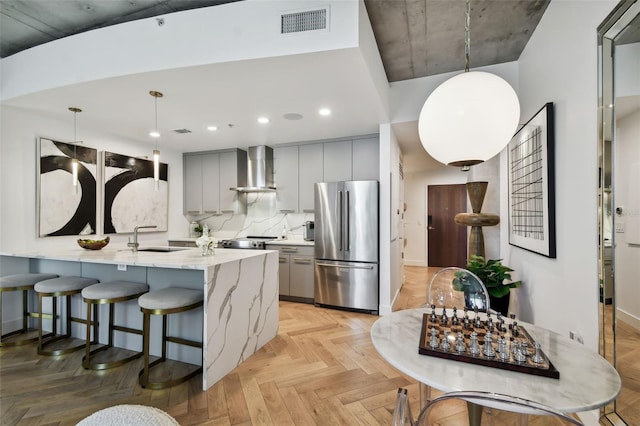 kitchen featuring gray cabinetry, stainless steel refrigerator, kitchen peninsula, pendant lighting, and wall chimney range hood