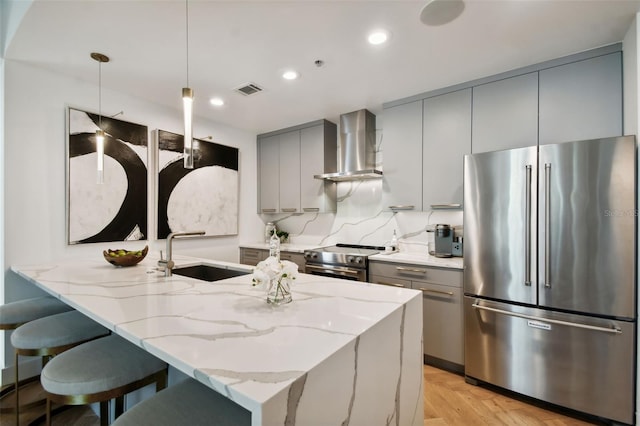 kitchen featuring sink, wall chimney range hood, appliances with stainless steel finishes, decorative light fixtures, and light stone countertops