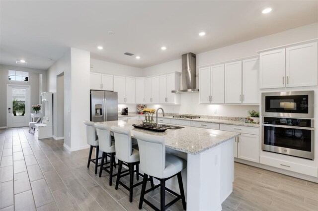 kitchen with white cabinets, a kitchen island with sink, stainless steel appliances, and wall chimney range hood