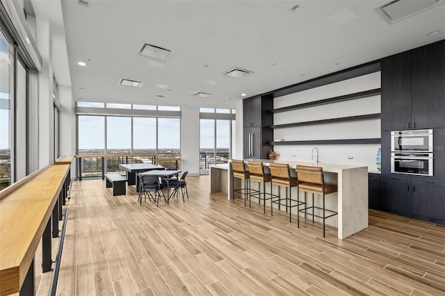 interior space with a breakfast bar area, expansive windows, a kitchen island with sink, stainless steel double oven, and light wood-type flooring