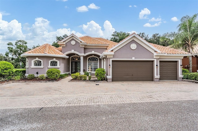 mediterranean / spanish-style home with a tile roof, decorative driveway, stucco siding, and an attached garage