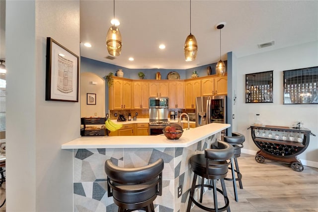 kitchen with decorative light fixtures, stainless steel appliances, light brown cabinetry, a breakfast bar area, and light hardwood / wood-style floors