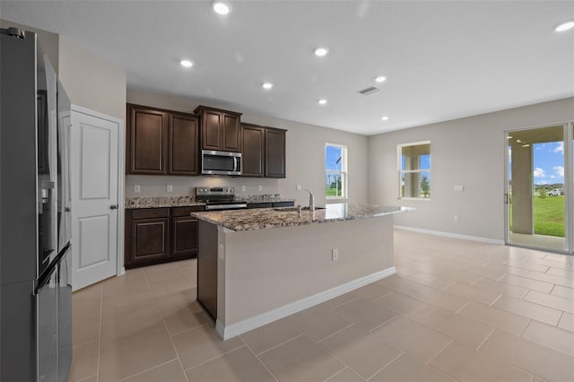 kitchen featuring appliances with stainless steel finishes, light tile patterned flooring, light stone countertops, dark brown cabinets, and a center island with sink