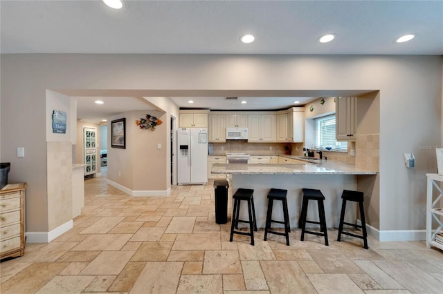 kitchen featuring kitchen peninsula, white appliances, cream cabinetry, a breakfast bar area, and decorative backsplash