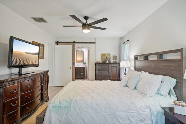 bedroom with connected bathroom, light wood-type flooring, a barn door, a textured ceiling, and ceiling fan