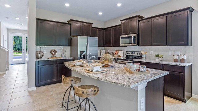 kitchen featuring dark brown cabinets, an island with sink, a kitchen bar, backsplash, and appliances with stainless steel finishes