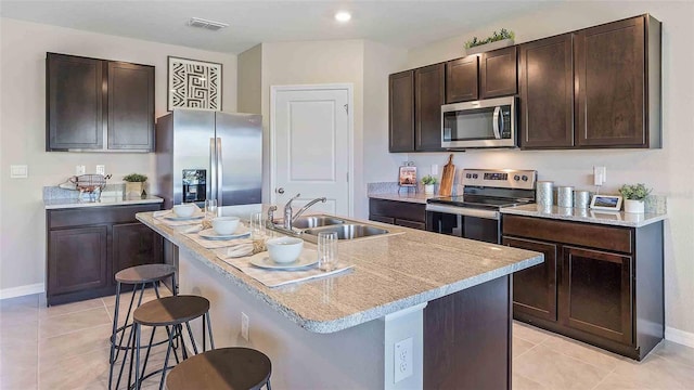 kitchen with light tile patterned flooring, an island with sink, stainless steel appliances, a kitchen bar, and dark brown cabinetry