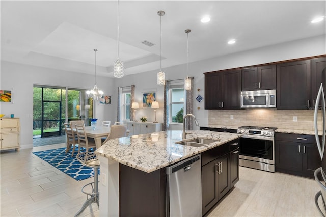 kitchen with pendant lighting, a kitchen island with sink, sink, appliances with stainless steel finishes, and a tray ceiling