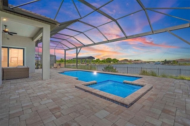 pool at dusk with a water view, ceiling fan, a lanai, an in ground hot tub, and a patio