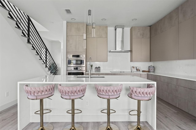 kitchen featuring wall chimney range hood, light hardwood / wood-style flooring, a kitchen bar, and a kitchen island with sink