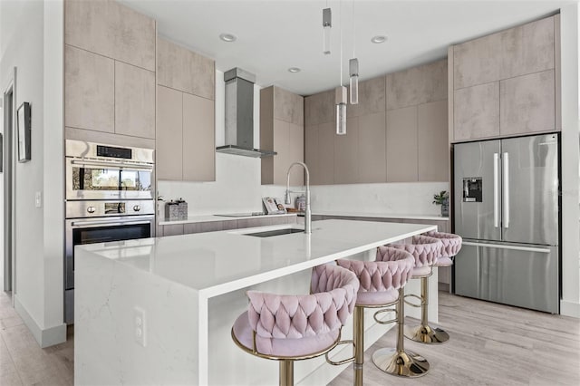 kitchen with wall chimney range hood, a center island with sink, a kitchen breakfast bar, stainless steel appliances, and decorative light fixtures