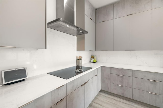 kitchen with black electric cooktop, light stone counters, backsplash, wall chimney exhaust hood, and gray cabinetry