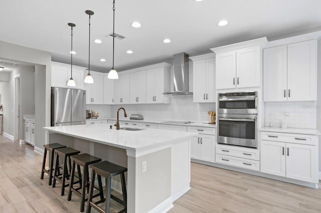 kitchen featuring sink, white cabinets, wall chimney exhaust hood, stainless steel appliances, and a center island with sink