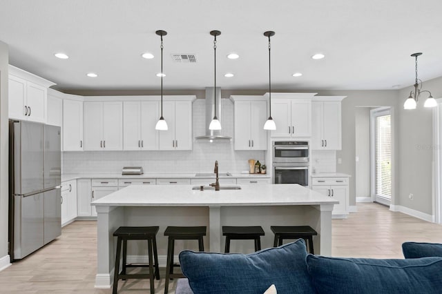 kitchen featuring sink, wall chimney range hood, a center island with sink, appliances with stainless steel finishes, and decorative light fixtures