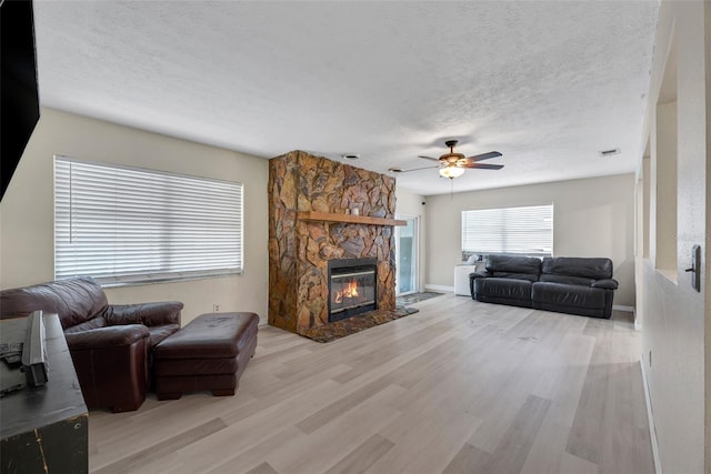 living room featuring ceiling fan, a stone fireplace, light hardwood / wood-style floors, and a textured ceiling