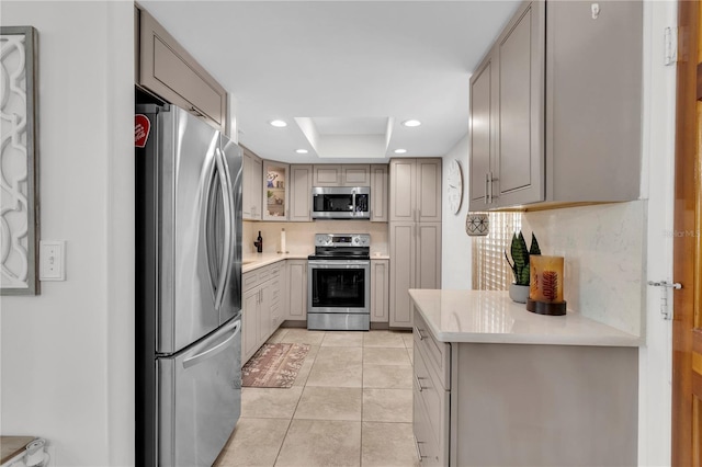 kitchen featuring gray cabinetry, a raised ceiling, light tile patterned floors, appliances with stainless steel finishes, and tasteful backsplash