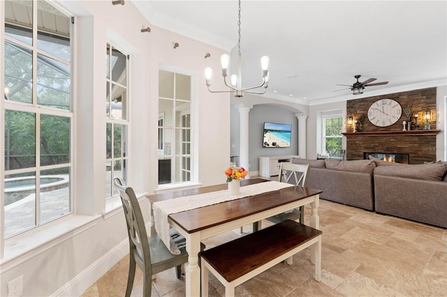 dining space featuring ceiling fan, a stone fireplace, baseboards, ornamental molding, and ornate columns