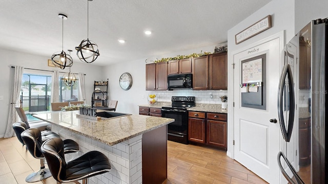 kitchen featuring light stone counters, a chandelier, a kitchen island with sink, black appliances, and decorative light fixtures