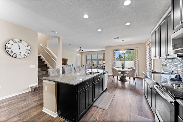 kitchen with a sink, a kitchen island with sink, baseboards, appliances with stainless steel finishes, and dark wood-style floors