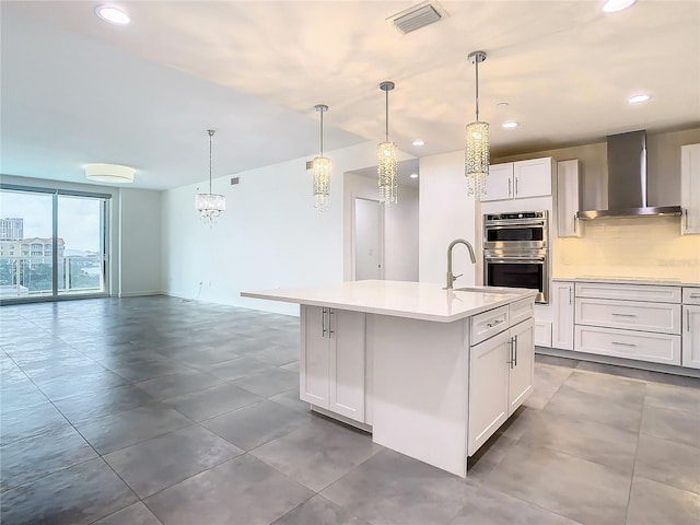 kitchen with a center island with sink, hanging light fixtures, white cabinets, double oven, and wall chimney exhaust hood