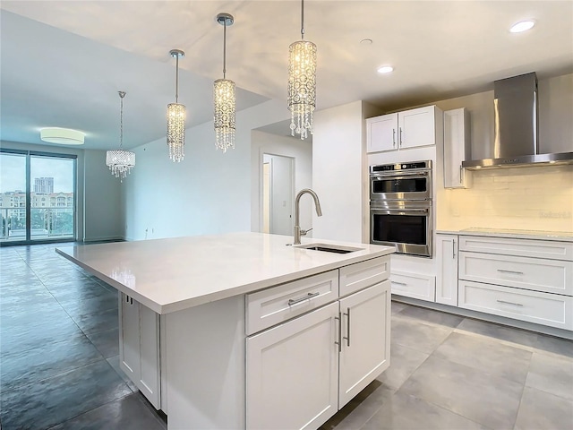 kitchen with a center island with sink, wall chimney exhaust hood, double oven, white cabinets, and sink