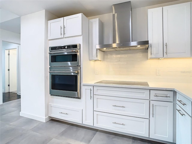 kitchen featuring backsplash, wall chimney range hood, black electric stovetop, white cabinetry, and stainless steel double oven