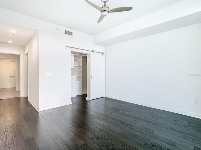 unfurnished bedroom with a barn door, ceiling fan, dark wood-type flooring, a closet, and a walk in closet