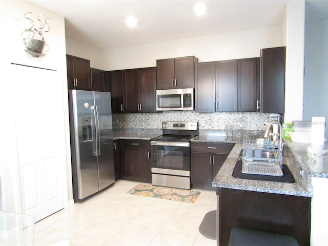 kitchen with dark brown cabinetry, sink, backsplash, stainless steel appliances, and light stone countertops