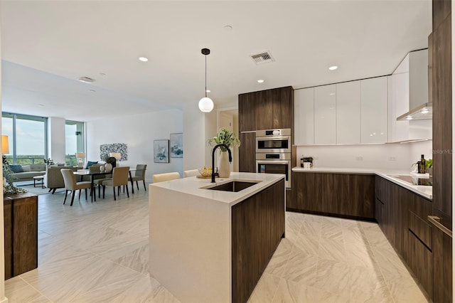 kitchen featuring stainless steel double oven, sink, pendant lighting, white cabinets, and an island with sink