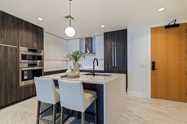 kitchen featuring wall chimney exhaust hood, stainless steel double oven, decorative light fixtures, a breakfast bar area, and an island with sink