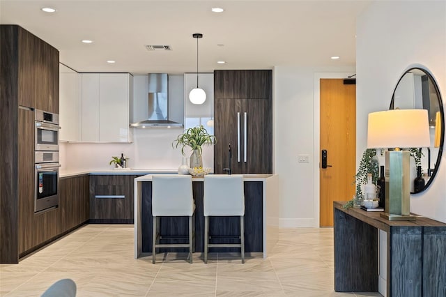 kitchen featuring a kitchen bar, dark brown cabinets, wall chimney range hood, decorative light fixtures, and white cabinetry