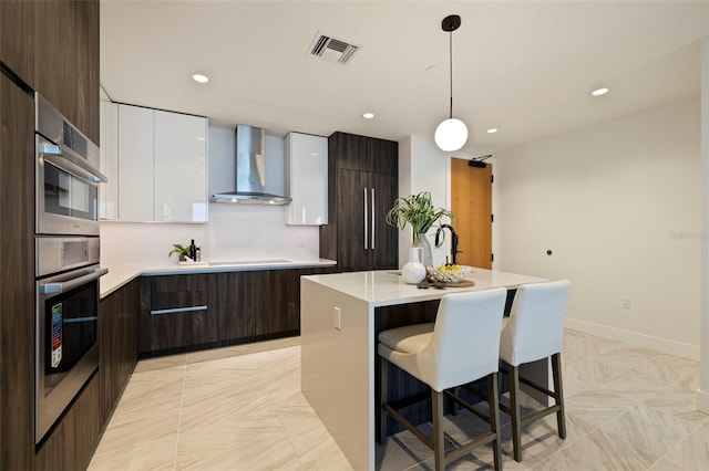 kitchen featuring dark brown cabinetry, wall chimney exhaust hood, hanging light fixtures, a center island with sink, and white cabinets