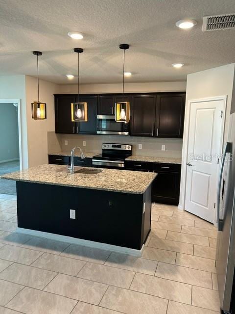 kitchen featuring stainless steel appliances, a sink, a textured ceiling, light stone countertops, and a center island with sink