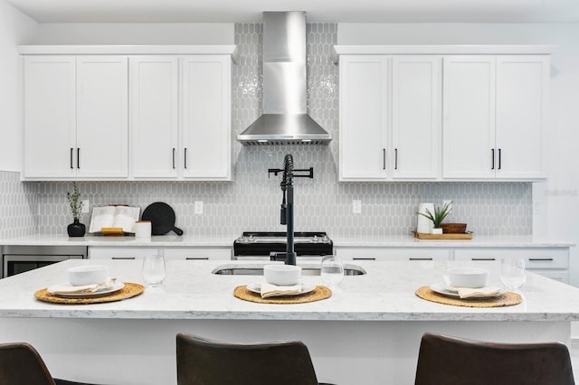 kitchen featuring wall chimney exhaust hood, white cabinetry, a center island with sink, and a breakfast bar area