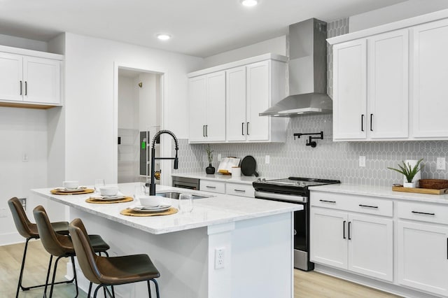 kitchen featuring white cabinets, wall chimney exhaust hood, stainless steel stove, a center island with sink, and sink