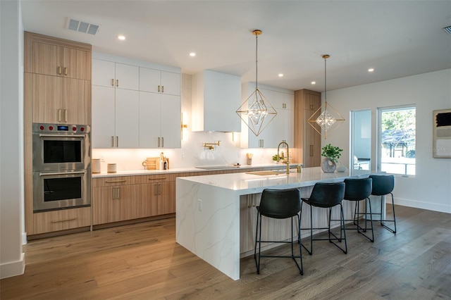 kitchen with white cabinetry, light hardwood / wood-style flooring, double oven, decorative light fixtures, and a kitchen island with sink