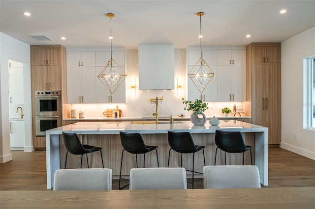 kitchen with hanging light fixtures, white cabinetry, double oven, and an island with sink