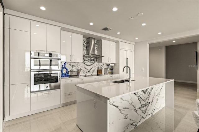 kitchen with wall chimney exhaust hood, a kitchen island with sink, sink, white cabinets, and double oven