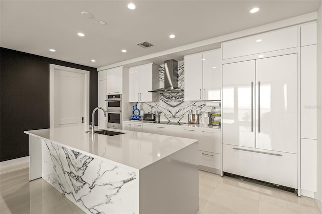 kitchen featuring wall chimney exhaust hood, paneled built in refrigerator, a center island with sink, white cabinets, and double oven