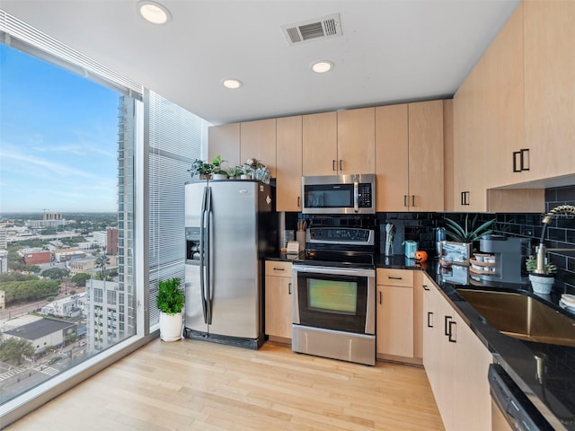 kitchen featuring light wood-type flooring, decorative backsplash, light brown cabinets, appliances with stainless steel finishes, and dark stone countertops