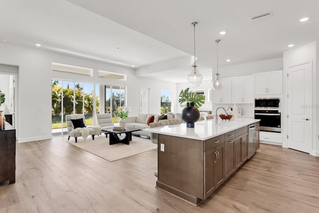 kitchen featuring white cabinetry, hanging light fixtures, light wood-type flooring, appliances with stainless steel finishes, and a kitchen island with sink