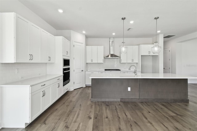 kitchen featuring white cabinetry, decorative light fixtures, a center island with sink, black oven, and wall chimney range hood