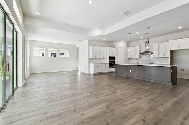 kitchen with hanging light fixtures, black oven, white cabinets, a kitchen island with sink, and wall chimney range hood