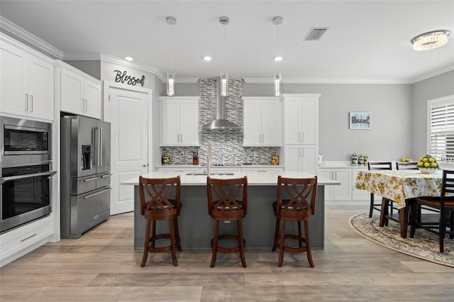 kitchen featuring wall chimney range hood, an island with sink, hanging light fixtures, white cabinetry, and stainless steel appliances