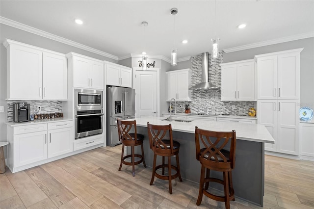 kitchen with appliances with stainless steel finishes, hanging light fixtures, wall chimney exhaust hood, white cabinets, and a center island with sink